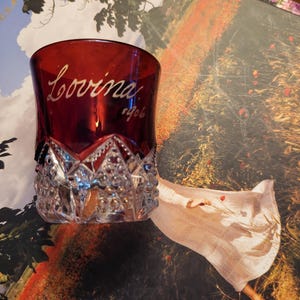 May include: A deep red glass tumbler featuring a clear, faceted crystal base with intricate geometric patterns. The upper portion of the glass is smooth, dark red, and displays the white cursive text "Lovina" followed by the number "1906" below it.