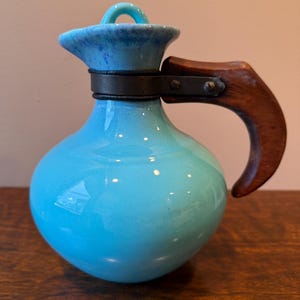 May include: A turquoise ceramic pitcher with a wooden handle and a dark metal band. The lid is a darker blue with a loop handle. The pitcher has a bulbous body and a spout.