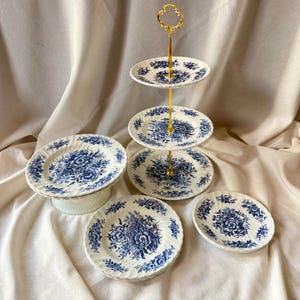 May include: A white and blue floral pattern tea set. The set includes a three-tiered serving stand with a gold handle, and several plates with a matching floral design. The plates are arranged on a white surface.