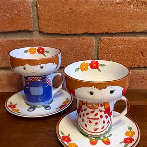 May include: Two ceramic cup and saucer sets featuring character designs. One cup depicts a figure in blue overalls, the other a figure in a strawberry-print apron. Both cups have wide rims with painted orange and red flowers and matching saucers.
