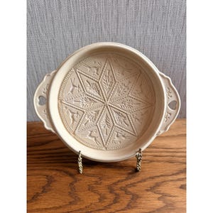 May include: A round, cream-colored ceramic baking dish with two side handles featuring heart-shaped cutouts. The interior base displays an embossed, intricate star pattern alternating with detailed depictions of leaping deer and pine trees.