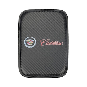 Puede incluir: Funda de asiento de coche rectangular negra con un patrón de fibra de carbono. El logotipo de Cadillac está en la funda, con la palabra "Cadillac" en escritura roja. La funda tiene un borde negro.