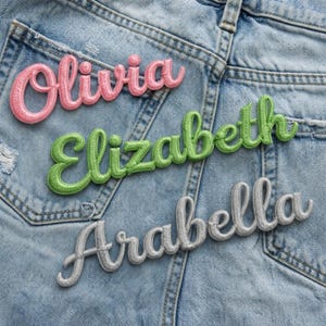 Könnte beinhalten: Gestickte Namensaufnäher in rosa, grün und silberner Schrift. Die Namen "Olivia", "Elizabeth" und "Arabella" sind auf einem hellblauen Jeanshintergrund abgebildet, wahrscheinlich eine Jeansjacke oder -tasche.