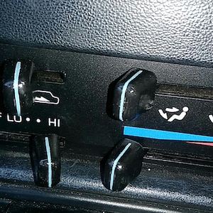 May include: Close-up of a car's dashboard with black control knobs. Each knob has a vertical light blue stripe. The dashboard has white icons and text, including "OFF", "LO", and "HI".