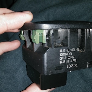 May include: A black electronic component with green clips, held in a hand. The component has the text "NC11 66 3508 02", "OMRON", "C8H-D102-02", "MADE IN JAPAN", and "1566D4" printed on it. The background is a dark, textured surface.