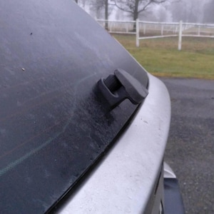May include: A close-up of the rear window of a silver car, featuring a black wiper blade. The window is slightly dirty, with visible defroster lines. The background shows a foggy, overcast day with a white fence.
