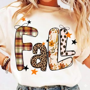 Könnte beinhalten: Ein cremefarbenes T-Shirt mit dem Wort Fall in großen, stilisierten Buchstaben. Jeder Buchstabe hat ein einzigartiges Muster: Schottenkaro, Leopardenmuster, Blumen und Punkte. Das Design ist mit kleinen schwarzen und orangen Sternen verziert.
