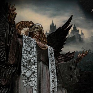May include: A figure wearing a gold filigree mask with hanging chains, a dark textured robe with white floral lace panels, and large black feathered wings. The figure has gold claw-like finger extensions and a circular gold halo-like headpiece.
