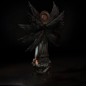 May include: A dark-toned figurine of a winged entity with multiple black feathered wings and a circular bronze-colored halo behind its head. The figure stands on a rocky base adorned with small, lit orange candles, set against a black background.