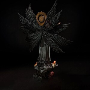 May include: A detailed figurine of a winged figure with multiple large black feathered wings and a circular gold halo behind the head. The figure wears a long dark garment and stands on a rocky base decorated with small lit candles and red accents.
