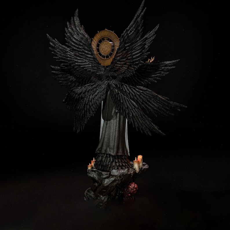 May include: A detailed figurine of a winged figure with multiple large black feathered wings and a circular gold halo behind the head. The figure wears a long dark garment and stands on a rocky base decorated with small lit candles and red accents.