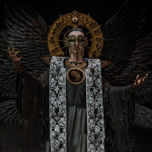 May include: A dark costume featuring large black feathered wings, a gold halo headpiece with a central eye, and a metallic plague doctor mask. The attire includes a black robe with a white patterned stole and a circular chest emblem with gold details.