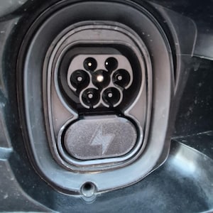 May include: Close-up of an electric vehicle charging port. The port is black with circular pin receptacles. A cover with a lightning bolt symbol is below the port. The surrounding area is dark blue.