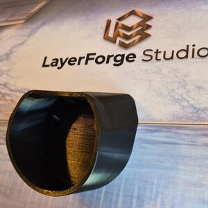 May include: A black, curved object with a hollow interior, possibly a 3D-printed component. The object is positioned against a backdrop featuring the text "LayerForge Studios" and a geometric logo. The lighting creates highlights and shadows on the curved surface.
