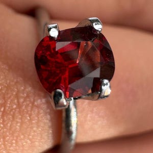 Op de afbeelding: Een close-up van een zilveren ring met een grote, ronde, dieprode edelsteen. De edelsteen is gefacetteerd, vangt het licht en vertoont een schitterende glans. De ring heeft een eenvoudige, gepolijste zilveren band.