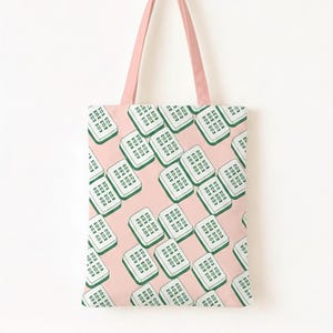 Peut inclure: Un sac fourre-tout rose avec un motif répétitif de carrés verts et blancs. Le sac a des anses roses et est suspendu à une cheville en bois sur un mur blanc. Le motif semble être un design stylisé.