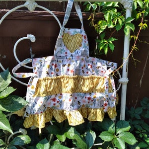 May include: A white apron with a yellow honeycomb pattern and a heart-shaped pocket. The apron has a ruffled bottom and is decorated with bees, flowers, and leaves.