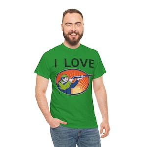 Op de afbeelding: Groen t-shirt met de tekst "I LOVE" boven een ovale afbeelding van een persoon die met een geweer mikt. De afbeelding is geplaatst tegen een opkomende zon. De persoon draagt een groene hoed en jas.