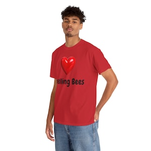 Op de afbeelding: Rood t-shirt met een groot, glanzend rood hart en de zwarte tekst "Spelling Bees". Het shirt met korte mouwen is effen rood van kleur. Het hart en de tekst zijn gecentreerd op de borst.