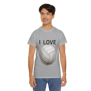 Op de afbeelding: Een lichtgrijs t-shirt met de tekst "I LOVE" boven een volleybalafbeelding. De volleybal is wit en heeft zwarte naden. De persoon draagt een blauwe spijkerbroek.