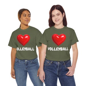 Op de afbeelding: Twee vrouwen dragen olijfgroene t-shirts met een groot rood hart en het woord "VOLLEYBALL" in het wit. De vrouwen dragen een blauwe jeans.