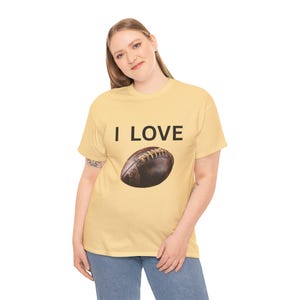 Op de afbeelding: Lichtgele t-shirt met de tekst "I LOVE" en een bruine voetbalafbeelding. De voetbal heeft witte veters. Het t-shirt wordt gedragen door een persoon die een blauwe spijkerbroek draagt.