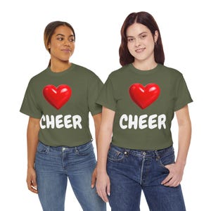 Op de afbeelding: Twee mensen dragen olijfgroene t-shirts met een groot rood hart en het woord 'CHEER' in witte letters. Ze dragen blauwe jeans. De afbeelding toont een casual stijl.