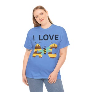 Op de afbeelding: Een lichtblauw t-shirt met de tekst "I LOVE ABC" in zwart. De letters A en C zijn ontworpen als bijen met gele en bruine strepen. Tussen de letters staat een blauwe vlinder.