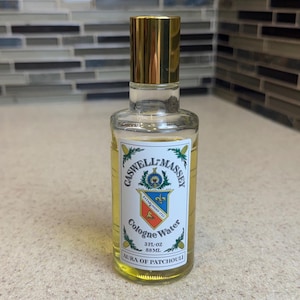 Peut inclure: Un flacon en verre transparent de Cologne Water Caswell-Massey, parfum Aura of Patchouli. Le flacon possède un bouchon doré et une étiquette blanche avec un blason bleu, rouge et or, indiquant 3 FL OZ et 88 ML. Le liquide est de couleur jaune pâle.