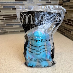 Può includere: Una statuetta blu di James P. Sullivan Sulley della Disney Pixar all'interno di un sacchetto di plastica trasparente McDonald's. La confezione presenta il logo McDonald's, il numero 6 e il testo che identifica il personaggio come James P. Sullivan Sulley in inglese, francese e spagnolo.