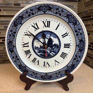 Op de afbeelding: Een witte keramische bordklok met een blauw en wit windmolenontwerp. De wijzerplaat heeft zwarte Romeinse cijfers en zwarte wijzers. De plaat wordt weergegeven op een bruine houten standaard.