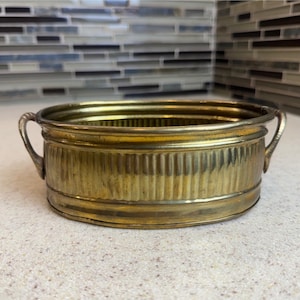 May include: An oval brass bowl featuring a ribbed, vertical texture along the exterior walls. The bowl has two small, curved handles attached to opposite sides and a smooth, flat rim. The metal surface displays a polished, metallic gold finish.