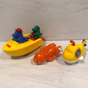 May include: A set of vintage TOMY bath toys. Features a yellow boat with blue and green characters, an orange sea creature, and a yellow submarine with a blue and white emblem.