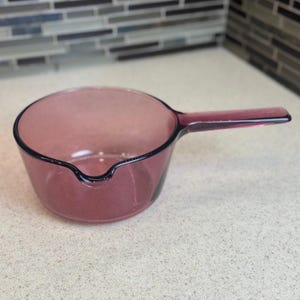 May include: A vintage, clear glass saucepan in a soft purple hue. The saucepan features a long, flat handle and a pouring spout. The rim is outlined with a dark purple trim. This kitchenware item is ideal for cooking and serving.