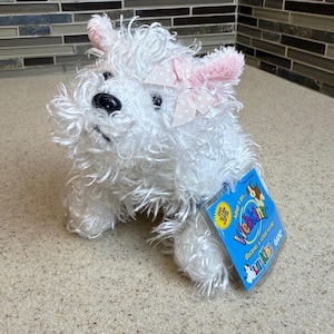 May include: A white plush West Highland Terrier dog with a pink bow and ears. The stuffed animal has a black nose and eyes. A Webkinz tag is attached to the dog.
