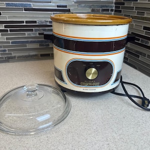 May include: A vintage Rival Crock-Pot slow cooker with a glass lid. The slow cooker is white with brown and blue accents, a gold-colored knob, and the words "RIVAL CROCK-POT SLOW-COOKER". The lid is clear glass.