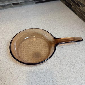 May include: An amber-coloured glass frying pan with a long handle. The pan is round with a dark rim, designed for cooking. The handle is an extension of the pan's body.
