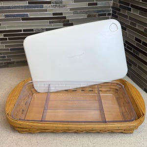 May include: A rectangular Longaberger basket with a clear plastic insert and a white lid. The basket is made of woven wood and has a clear plastic insert with three compartments. The white lid has a handle and the Longaberger logo.