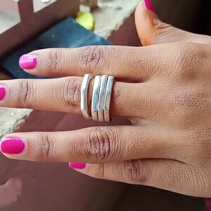 Könnte beinhalten: Ein Satz von vier silbernen Ringen mit rechteckigem Design ist auf einem Finger gestapelt. Die Ringe sind glatt und glänzend und reflektieren das Licht. Die Fingernägel sind hellrosa lackiert. Der Hintergrund ist eine verschwommene Werkbank.