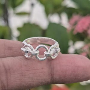 Könnte beinhalten: Ein silberner Ring mit einem Kettenglied-Design. Der Ring hat drei miteinander verbundene Kettenglieder, die ein einzigartiges und stilvolles Accessoire bilden. Der Ring wird auf einem Finger mit einem verschwommenen grünen und rosa Hintergrund gezeigt.