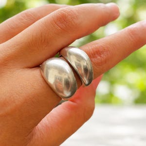 Könnte beinhalten: Zwei silberne Ringe mit glattem, abgerundetem Design. Die Ringe werden an einem Finger getragen, reflektieren das Licht und wirken glänzend. Die Ringe haben eine minimalistische, moderne Ästhetik.