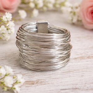 Könnte beinhalten: Ein silberfarbener Ring, der aus zahlreichen dünnen Drähten besteht, die eng miteinander gewickelt sind. Der Ring hat einen rechteckigen Verschluss und ist vor einem verschwommenen Hintergrund aus weißen Blumen und einer rosa Rose platziert. Ein Schmuckstück.