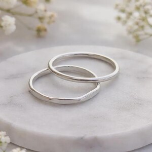 Könnte beinhalten: Zwei silberne Ringe, einer mit glattem Band und der andere mit facettiertem Design, sind auf einer weißen Marmoroberfläche gestapelt. Die Ringe stehen im Mittelpunkt, mit weichen, verschwommenen weißen Blumen im Hintergrund.