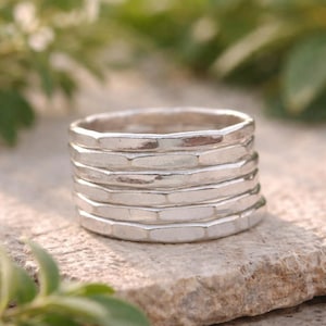 Könnte beinhalten: Ein Stapel von sechs silbernen Ringen mit einem strukturierten, facettierten Design. Die Ringe sind übereinander gestapelt, wodurch ein geschichteter Effekt entsteht. Die Ringe bestehen aus einem glänzenden, metallischen Material und werden auf einer Steinoberfläche präsentiert.