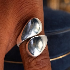 Könnte beinhalten: Ein silberner Ring mit einem einzigartigen Design. Der Ring hat zwei tropfenförmige Elemente, die den Finger umschließen. Der Ring besteht aus einem glänzenden, reflektierenden Metall.