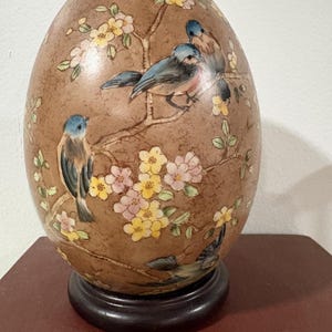 May include: A decorative egg-shaped ornament with a brown textured surface. The egg features a painted design of blue birds perched on flowering branches with pink and yellow blossoms. The egg sits on a dark brown wooden base.