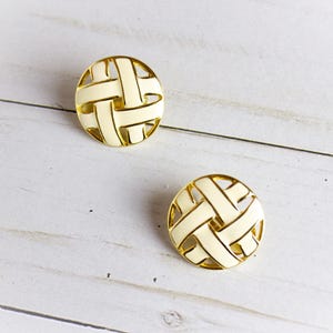 May include: Two round, cream-colored earrings with a woven design and gold-tone trim. The earrings are set against a white wooden background. The earrings are a classic accessory.