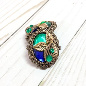 May include: A pair of oval-shaped earrings with a gold-tone metal setting. The earrings feature a blue and green gem with gold leaf accents.