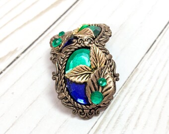 May include: A pair of oval-shaped earrings with a gold-tone metal setting. The earrings feature a blue and green gem with gold leaf accents.