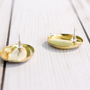 May include: Two gold-colored, oval-shaped stud earrings with silver posts. The earrings have a shiny, reflective surface. The earrings are displayed on a white wooden surface.